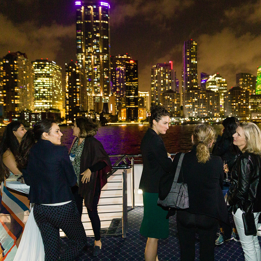 Many women conversing at an event by a river at night with brightly lit skyscrapers in the background.