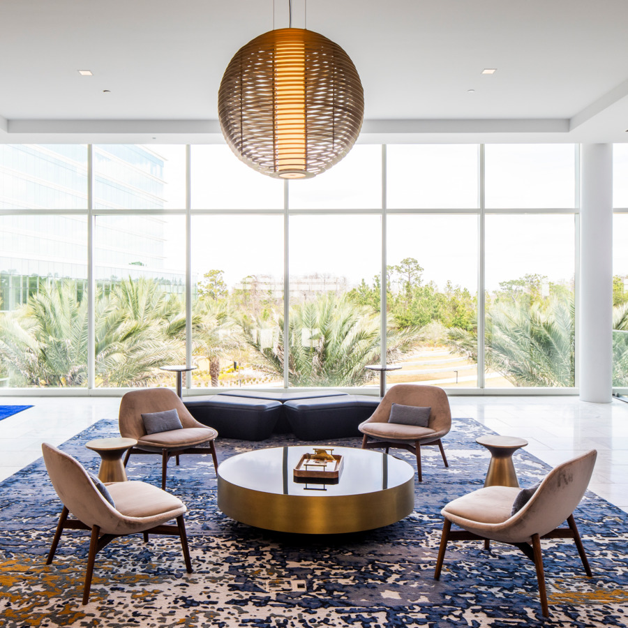 Brass coffee table holding a tray surrounded by 4 lounge chairs on a speckled rug with leather benches and a large window overlooking palm trees in the background